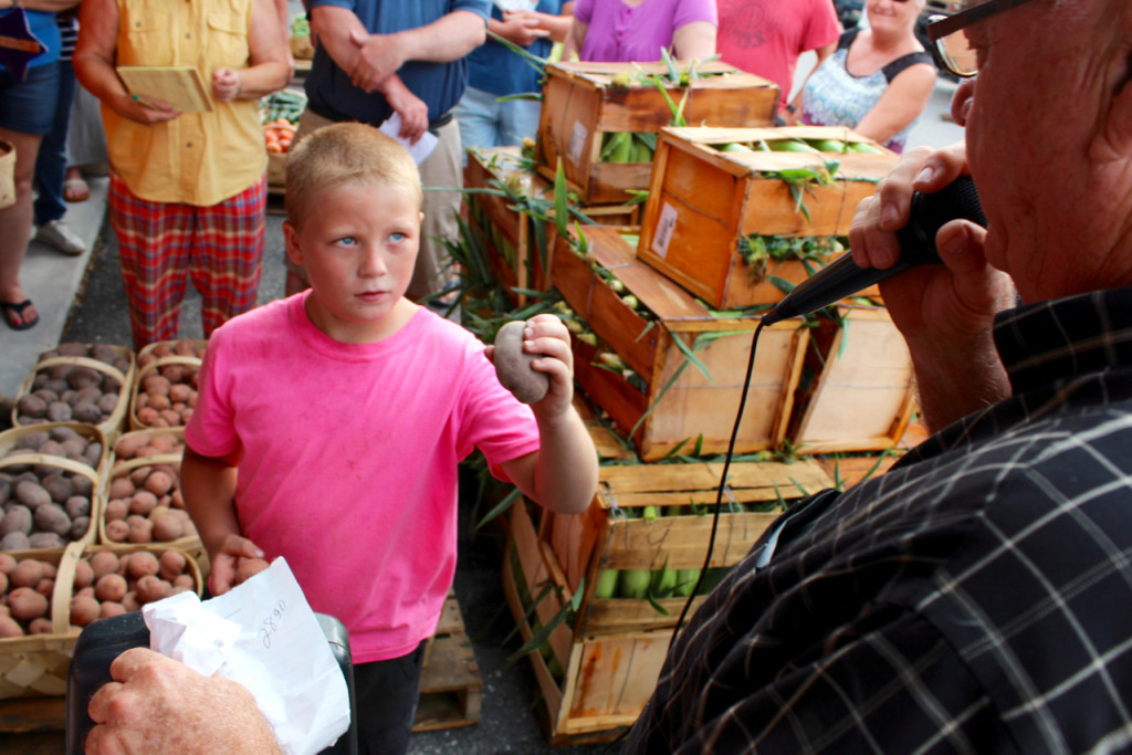 While auctioneer Gene Daniels, microphone in hand, works at a recent produce auction at the Virginia Beach Farmers Market, he has a little help from Alex Huban, 8, who shows off the produce up for bids. [John-Henry Doucette/The Princess Anne Independent News]