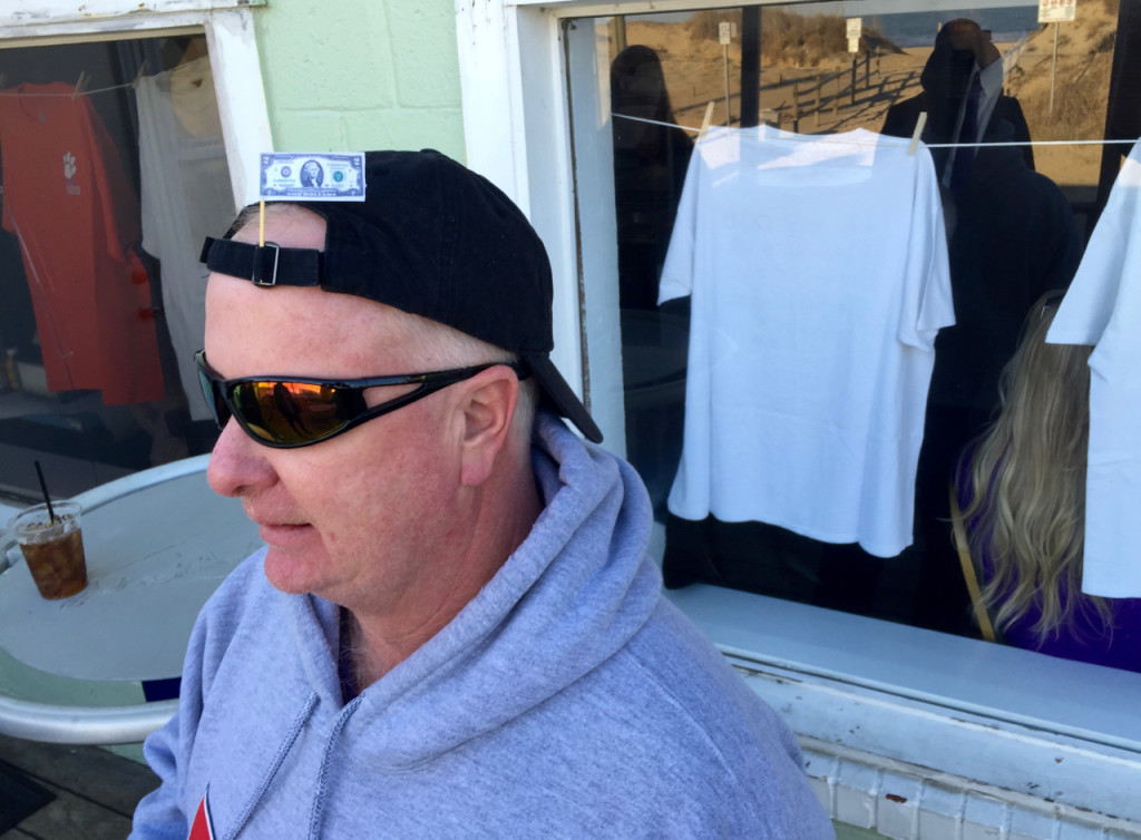 Chuck Mullis wears a decorative $2 bill in his ball cap during the gathering to celebrate Jones. In the window are some of Jones’ tees, which were on clothes lines on the walls of the restaurant. [John-Henry Doucette/The Princess Anne Independent News]