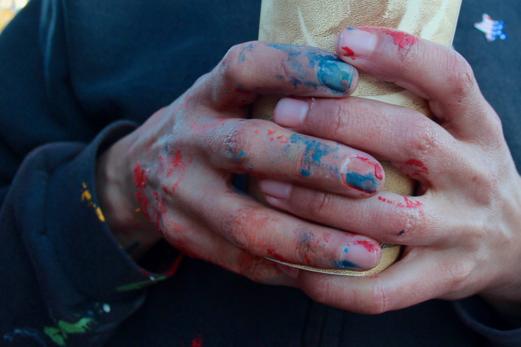 The artist’s hands, wrapped around a coffee cup, show remnants of the paint. The work will remain as long as the paint holds up to traffic. [John-Henry Doucette/The Princess Anne Independent News]